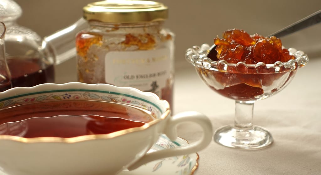 Photo of a glass compotière filled with marmalade, with a jar of Fortnum & Mason marmalade in the background and an elegant cup of tea in the foreground, capturing a refined tea-time setting.