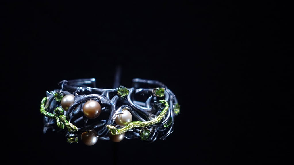 Photo of a contemporary bracelet featuring several pearls set in a grey metal mount, with bright green serpents intricately coiling around the structure, creating a bold and artistic design.
