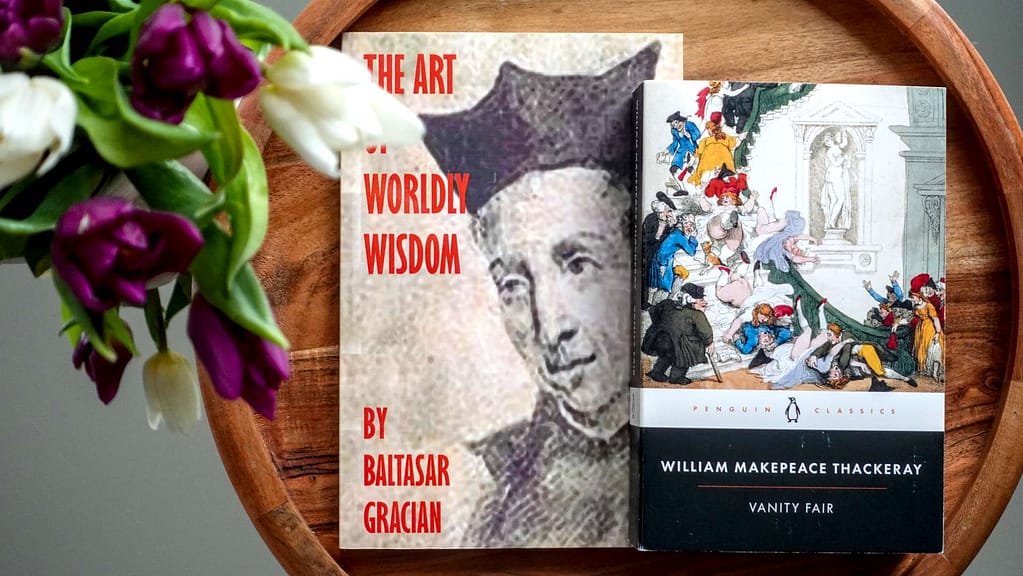 A flatlay of Vanity Fair by Thackeray and The Art of Worldly Wisdom by Baltasar Gracián with pink tulips, representing the intersection of modern etiquette and literature.