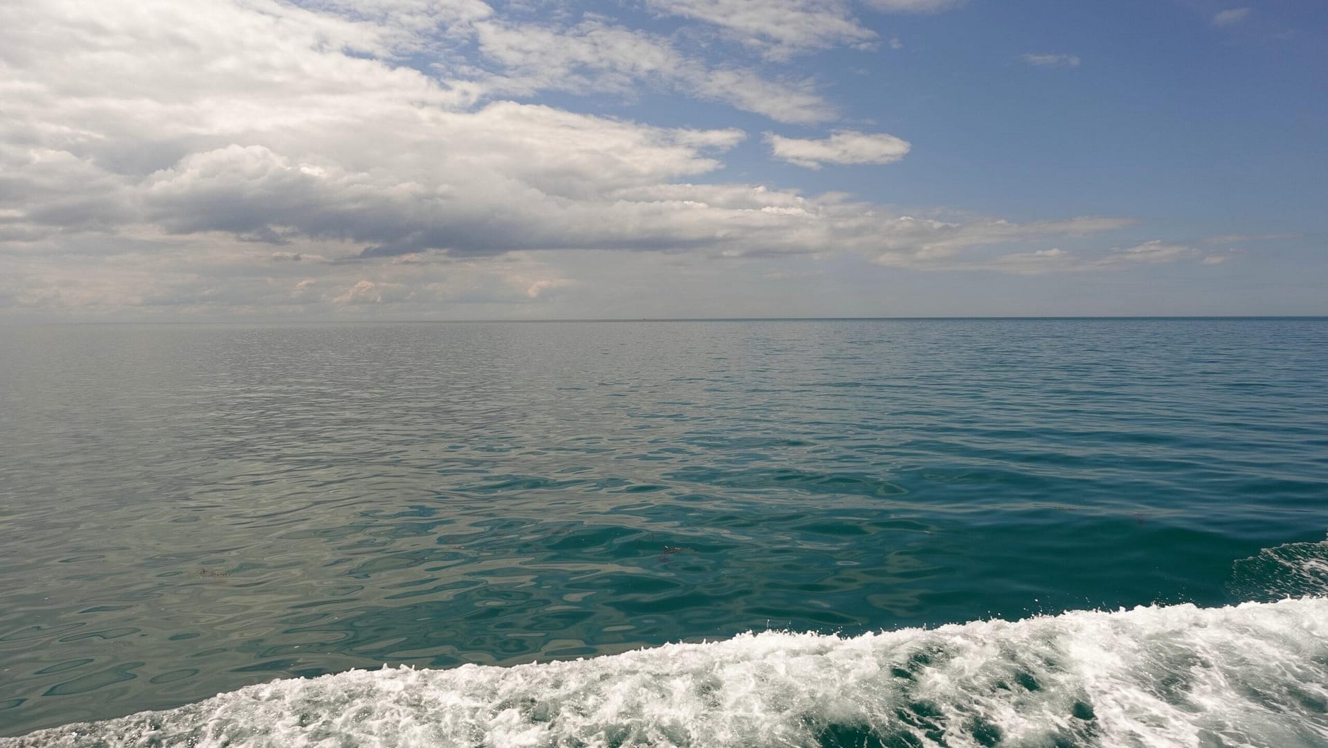 sea scape with white clouds in the sky, a dark green-blue sea and a wave of foam from the boat in the foreground