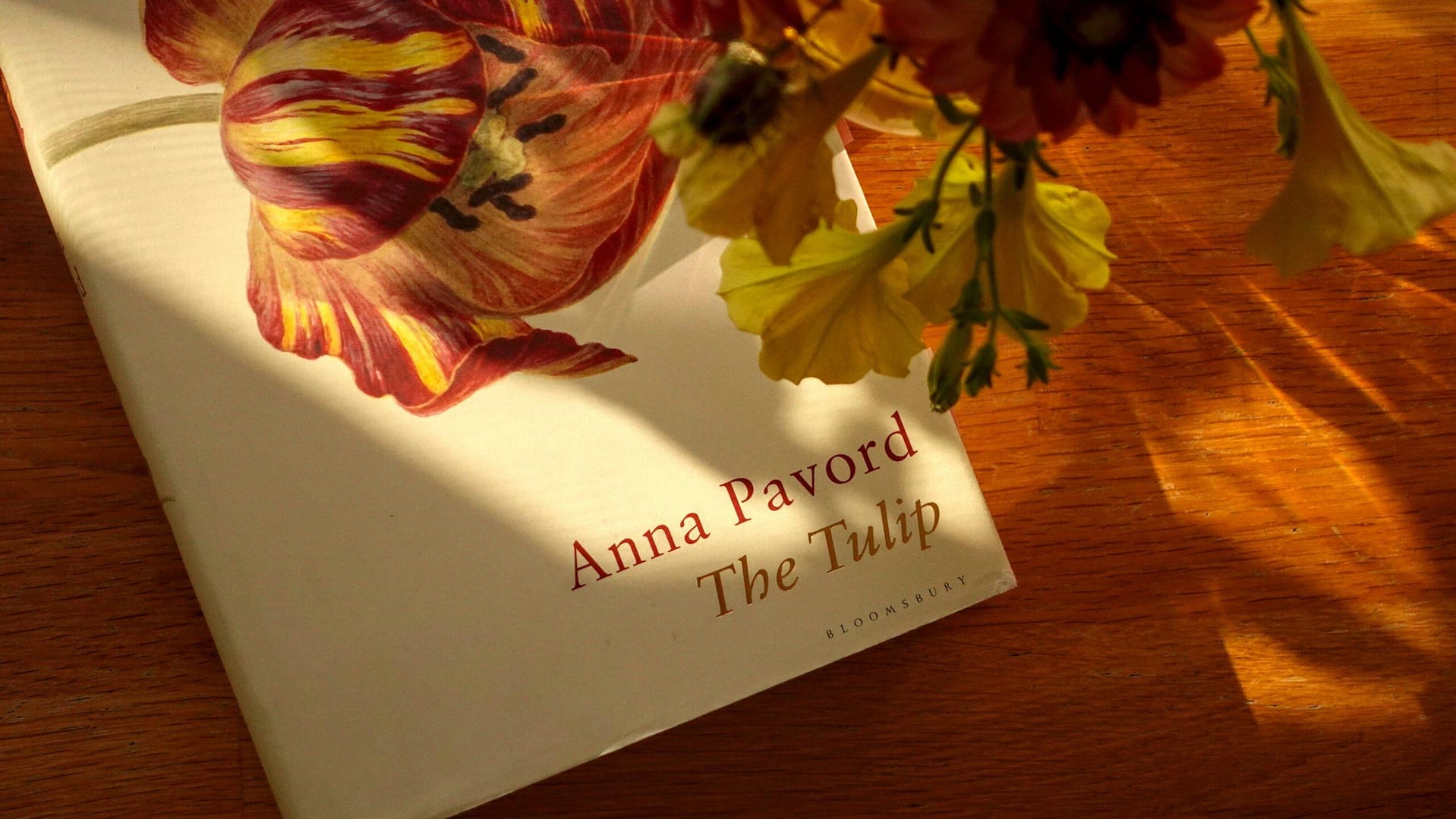 a copy of the book The Tulip by Anna Pavord with a bouquet of yellow petunias and dahlias on a sun lit wooden table